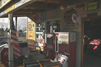 A rustic scene featuring various vintage advertisements and memorabilia. The area is decorated with old Coca-Cola vending machines, an American flag, retro metal signs from brands like Texaco and Mobil, and older automotive memorabilia. A wooden structure provides partial shade, and there are stacks of items such as records or books, contributing to a nostalgic atmosphere.