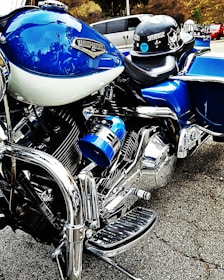 A close-up view of a blue and chrome Harley-Davidson motorcycle, emphasizing the intricate detailing of the engine and polished metal finishes. A helmet with stickers is resting on the seat. In the background, a blurred street setting with vehicles is visible.