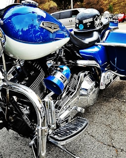 A close-up view of a blue and chrome Harley-Davidson motorcycle, emphasizing the intricate detailing of the engine and polished metal finishes. A helmet with stickers is resting on the seat. In the background, a blurred street setting with vehicles is visible.