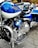A close-up view of a blue and chrome Harley-Davidson motorcycle, emphasizing the intricate detailing of the engine and polished metal finishes. A helmet with stickers is resting on the seat. In the background, a blurred street setting with vehicles is visible.