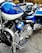 A close-up view of a blue and chrome Harley-Davidson motorcycle, emphasizing the intricate detailing of the engine and polished metal finishes. A helmet with stickers is resting on the seat. In the background, a blurred street setting with vehicles is visible.
