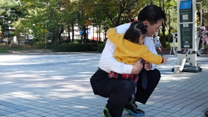 A person squatting while holding a child in a park setting with exercise equipment and trees in the background. The child is wearing a yellow sweater and has their arms wrapped around the adult.