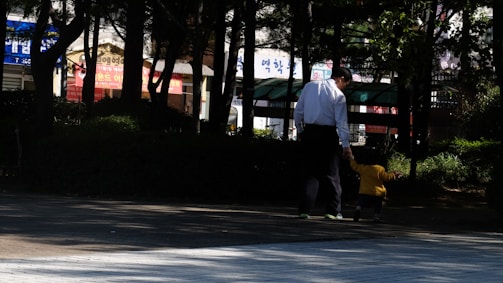 A father and child wearing matching donko t-shirts, walking hand in hand in a sunlit urban park.