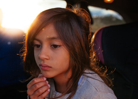 A young child with long hair sits in a car, appearing deep in thought or contemplation. The sunlight coming from behind adds a warm glow to the scene, partially illuminating the child's face and highlighting their pensive expression. The interior of the car is dimly lit except for the sunlight streaming in.