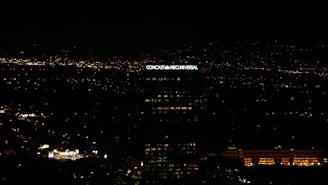A tall building with the illuminated sign of a well-known media company stands against a backdrop of a cityscape at night. Numerous small lights are scattered across the landscape, indicating urban activity in the distance.
