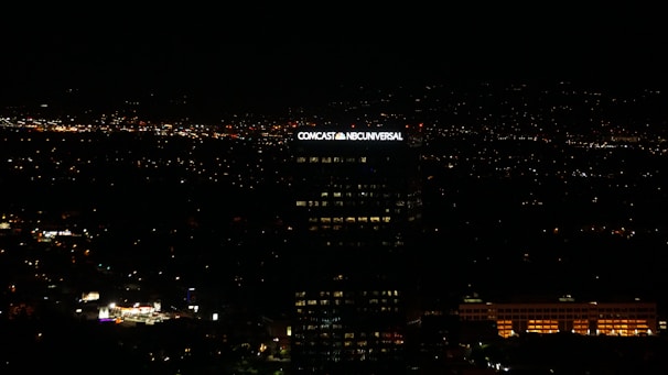 A tall building with the illuminated sign of a well-known media company stands against a backdrop of a cityscape at night. Numerous small lights are scattered across the landscape, indicating urban activity in the distance.