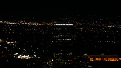 A tall building with the illuminated sign of a well-known media company stands against a backdrop of a cityscape at night. Numerous small lights are scattered across the landscape, indicating urban activity in the distance.