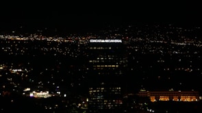 A tall building with the illuminated sign of a well-known media company stands against a backdrop of a cityscape at night. Numerous small lights are scattered across the landscape, indicating urban activity in the distance.