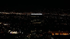 A tall building with the illuminated sign of a well-known media company stands against a backdrop of a cityscape at night. Numerous small lights are scattered across the landscape, indicating urban activity in the distance.