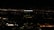 A tall building with the illuminated sign of a well-known media company stands against a backdrop of a cityscape at night. Numerous small lights are scattered across the landscape, indicating urban activity in the distance.