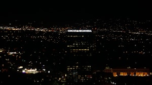 A tall building with the illuminated sign of a well-known media company stands against a backdrop of a cityscape at night. Numerous small lights are scattered across the landscape, indicating urban activity in the distance.