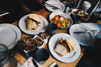 A wooden table is set with various plates of food, including sandwiches and a dish with nachos and toppings. Several drinks are visible, including a disposable coffee cup. Utensils and napkins are arranged neatly, and there's a small plant decoration on the table.