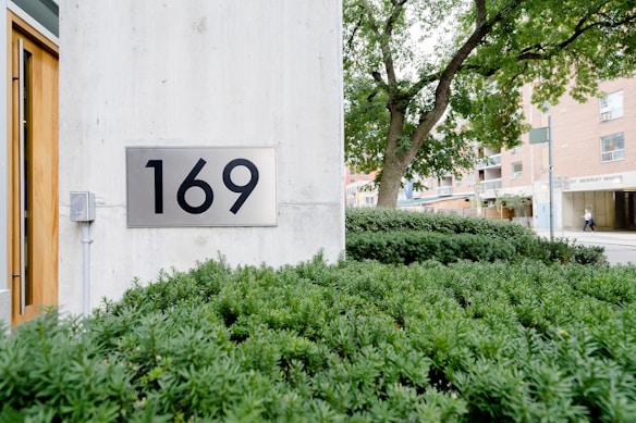 A concrete building wall with a large metal sign displaying the number 169. In the foreground, lush green shrubs are neatly arranged, and a tree with dense foliage stands next to the building. The background shows an urban street scene with a building labeled 'BEVERLY MANOR.'