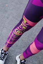 A person is wearing colorful athletic leggings featuring a mix of purple, pink, and yellow patterns and text. The lower legs are visible, along with black and white sneakers standing on an asphalt surface.