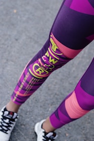A person is wearing colorful athletic leggings featuring a mix of purple, pink, and yellow patterns and text. The lower legs are visible, along with black and white sneakers standing on an asphalt surface.