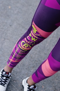 A person is wearing colorful athletic leggings featuring a mix of purple, pink, and yellow patterns and text. The lower legs are visible, along with black and white sneakers standing on an asphalt surface.