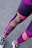 A person is wearing colorful athletic leggings featuring a mix of purple, pink, and yellow patterns and text. The lower legs are visible, along with black and white sneakers standing on an asphalt surface.