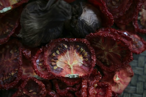 Slices of dried tomatoes with wrinkled, red skin and visible seeds arranged closely together. Some slices exhibit a dark, almost blackened appearance indicating a higher level of dehydration.