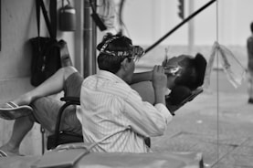 A man is sitting in a chair wearing glasses with magnifying lenses, working on the ear of another person reclining in a makeshift outdoor salon. The scene captures a moment of personal grooming in a street setting.