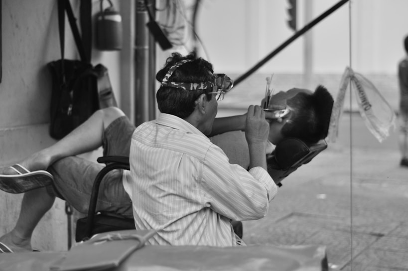 A man is sitting in a chair wearing glasses with magnifying lenses, working on the ear of another person reclining in a makeshift outdoor salon. The scene captures a moment of personal grooming in a street setting.