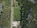 An aerial view of a rectangular sports field surrounded by dense green trees. A road runs parallel to one side, with clear shadows cast by tall structures beside the field. The grass is well-maintained and vibrant, with a small building or structure visible at one end of the field.