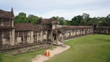 A historical stone structure with intricate carvings standing amidst lush green surroundings. Two people in bright orange and yellow traditional attire walk along a path beside the building. The architecture features tall, weathered walls and a partially ruined archway.