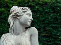 A marble statue of a woman depicted in a classical style, featuring detailed hair and delicate facial features, set against a lush green foliage background.