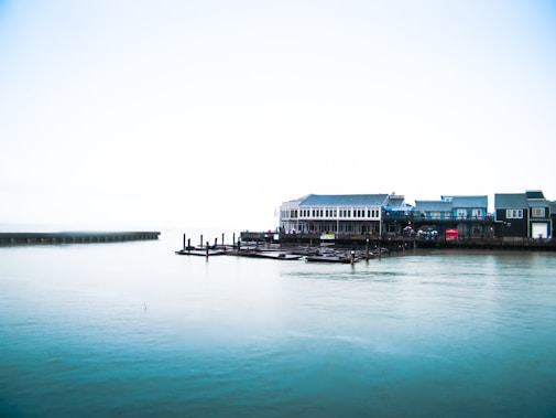 A waterfront scene with a large building complex situated near the edge of a calm body of water. Several docks extend into the water, and the sky is bright and slightly overcast, creating a serene atmosphere. The buildings have a modern architectural style with numerous windows.