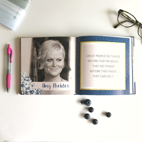 An open book displays a black and white photograph of a woman on the left page, surrounded by floral illustrations and her name written below. The right page features an inspirational quote set against a blue background with white speckles. On the table around the book are a pair of glasses, a pink pen, some blueberries, and the edge of a magazine.