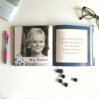 An open book displays a black and white photograph of a woman on the left page, surrounded by floral illustrations and her name written below. The right page features an inspirational quote set against a blue background with white speckles. On the table around the book are a pair of glasses, a pink pen, some blueberries, and the edge of a magazine.