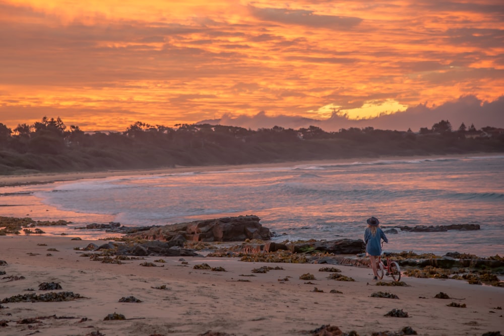 girl with bike on beach beside body of water under sunset