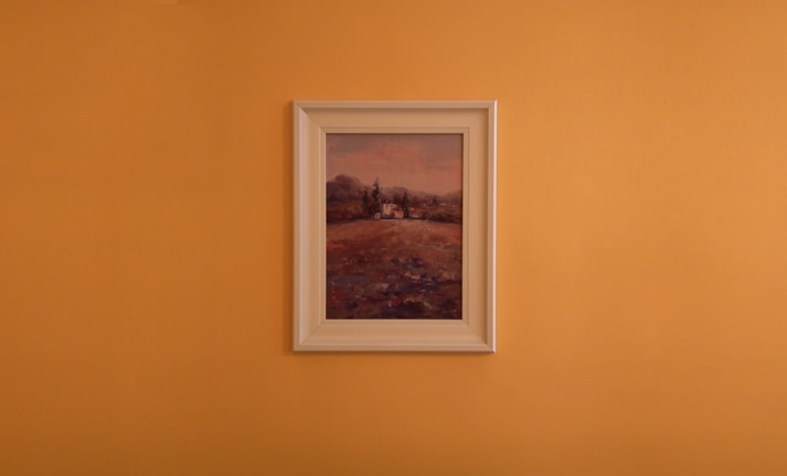 A framed landscape painting is centered against a plain orange wall. The artwork features a countryside scene with earthy tones, displaying a field leading up to some trees and distant hills under a muted sky.