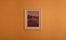 A framed landscape painting is centered against a plain orange wall. The artwork features a countryside scene with earthy tones, displaying a field leading up to some trees and distant hills under a muted sky.