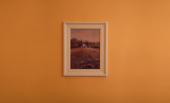 A framed landscape painting is centered against a plain orange wall. The artwork features a countryside scene with earthy tones, displaying a field leading up to some trees and distant hills under a muted sky.