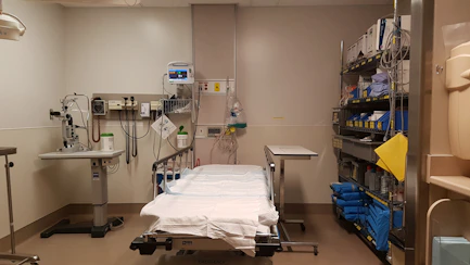 A hospital or medical examination room containing a bed with white sheets surrounded by medical equipment, such as monitors and diagnostic tools. There are shelves filled with supplies and medical instruments on the right side of the room.