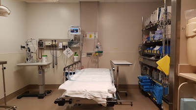 A hospital or medical examination room containing a bed with white sheets surrounded by medical equipment, such as monitors and diagnostic tools. There are shelves filled with supplies and medical instruments on the right side of the room.