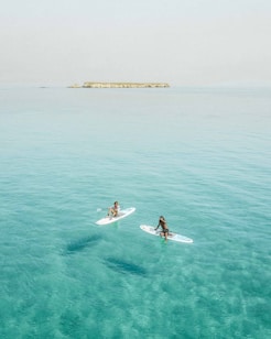 Two individuals are paddleboarding on calm, clear turquoise waters with an island visible in the distance. The scene is serene and peaceful, suggesting a leisurely day on the water.