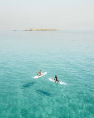 Two individuals are paddleboarding on calm, clear turquoise waters with an island visible in the distance. The scene is serene and peaceful, suggesting a leisurely day on the water.