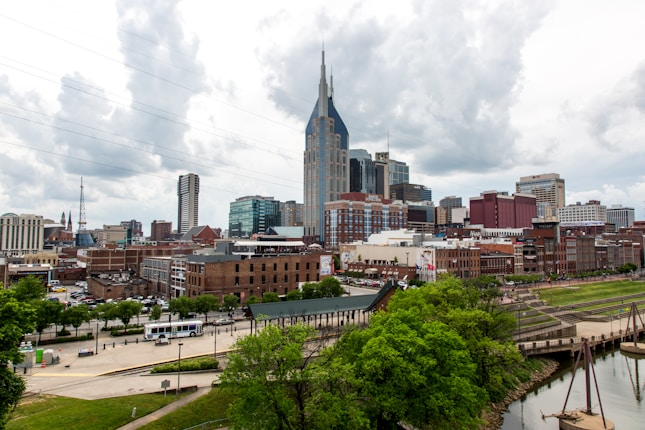 A cityscape featuring a skyline with tall modern skyscrapers and some historical brick buildings. The iconic AT&T Building is prominent, with its distinctive twin spires. The foreground shows green trees and a river, while the background is filled with thick clouds.