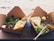 Two cardboard food containers are placed on a wooden surface. Each container holds a meal with grilled fish topped with capers and herbs. Surrounding the fish are fresh greens and a slice of lemon. Both meals are accompanied by folded flatbread and a small serving of some white condiment. Wooden utensils are placed in and around the containers. In the background, there is a sandy beach area.