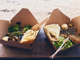 Two cardboard food containers are placed on a wooden surface. Each container holds a meal with grilled fish topped with capers and herbs. Surrounding the fish are fresh greens and a slice of lemon. Both meals are accompanied by folded flatbread and a small serving of some white condiment. Wooden utensils are placed in and around the containers. In the background, there is a sandy beach area.