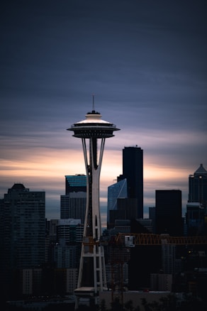 The image depicts a cityscape with a prominent tower in the foreground, likely the Space Needle, set against a backdrop of modern skyscrapers. The sky is overcast with a mix of dark clouds and a hint of sunset or sunrise light on the horizon, creating a dramatic contrast.