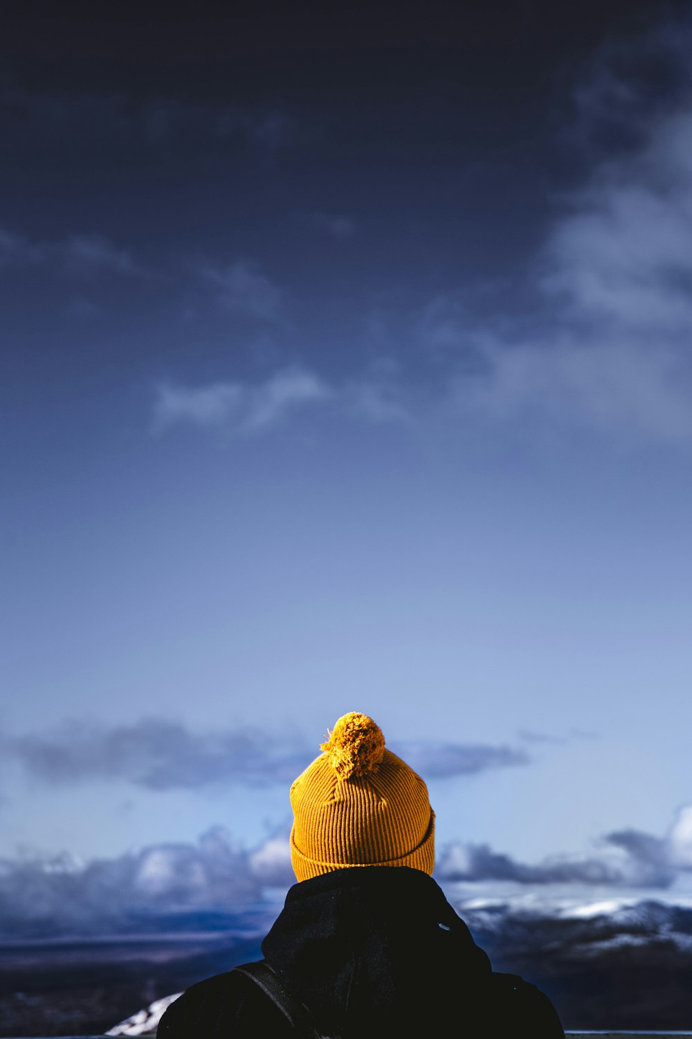 person wearing brown bobble hat facing cloudy skies