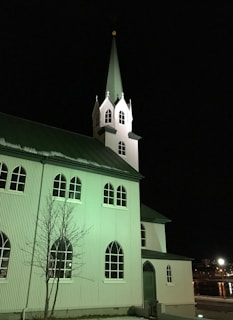A white church building with a tall, pointed steeple is illuminated at night. The structure features arched windows and a corrugated metal surface. The lighting casts a greenish hue over the facade. A barren tree stands in front of the building. In the background, city lights are reflected on a body of water.