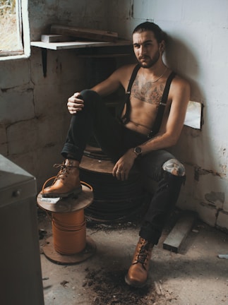 A man with a tattooed chest is sitting on a wooden reel in a dimly lit, rustic room. He is wearing suspenders, dark pants with a ripped knee, and brown work boots. The room appears to be industrial with bare, aged walls and a dirt floor.