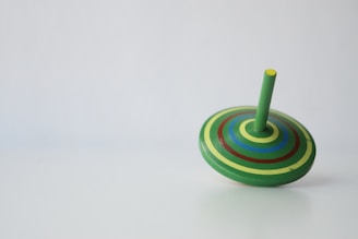 A colorful spinning top lies on a plain surface. The top is adorned with concentric circles in shades of green, yellow, blue, and red, with a green stem protruding from the center.