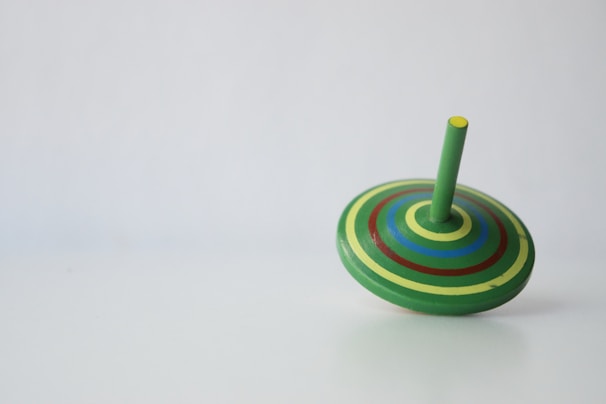 A colorful spinning top lies on a plain surface. The top is adorned with concentric circles in shades of green, yellow, blue, and red, with a green stem protruding from the center.
