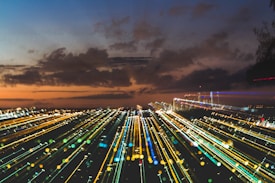 A dynamic cityscape at dusk with streaks of colorful lights creating a sense of motion, against a backdrop of dark clouds and an orange-tinged sky.