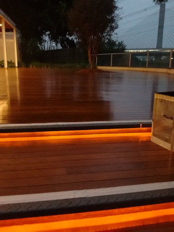 A wooden deck is illuminated by warm orange lights embedded in the stairs. The background features trees, a modern bridge with suspension cables, and a railing.