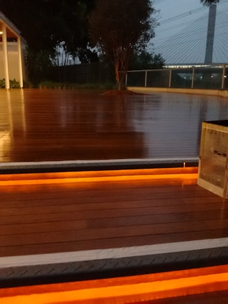 A wooden deck is illuminated by warm orange lights embedded in the stairs. The background features trees, a modern bridge with suspension cables, and a railing.